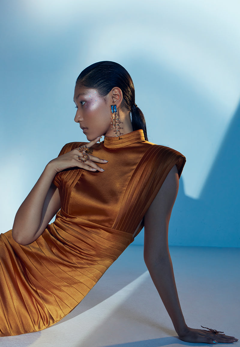 Designed to create a statement, this bodycon bandage dress in rustic gold is cut and constructed using lush satin. The sleeveless midi dress features criss-cross bandage draping with exaggerated shoulder padding and a pleated turtle neck. It has a zip closure at the center back and a flap opening at the back hem for easy movement. It is the perfect dress for your next formal dinner, date night, red carpet or a soiree. Pair yours with heeled sandals or pumps and long gold earrings to complete the look.