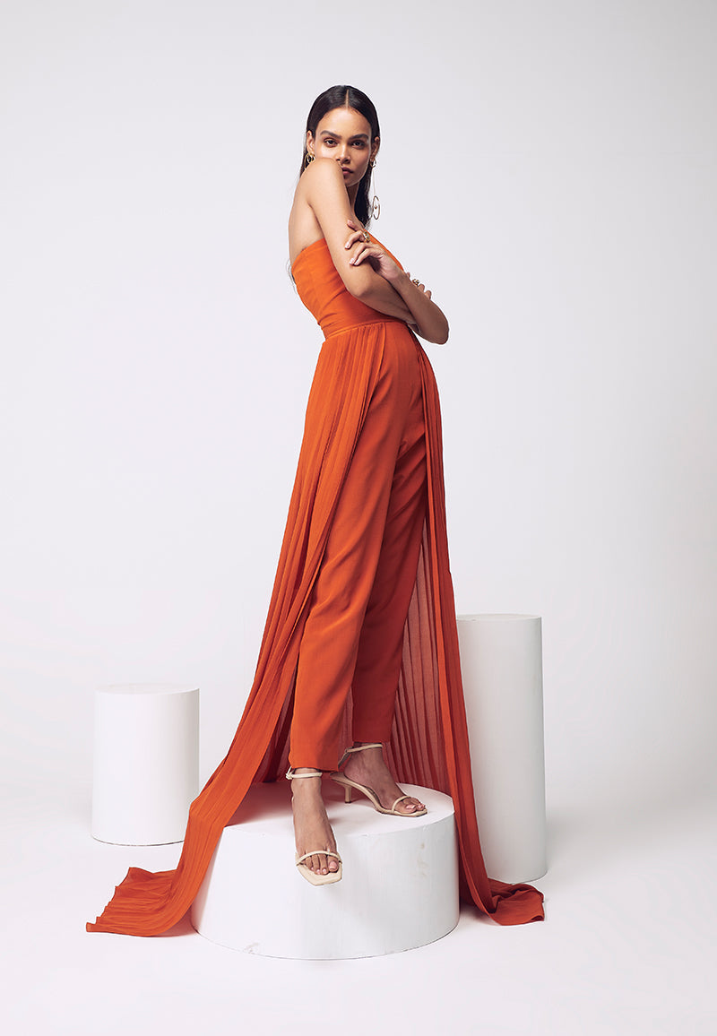 New style figure-hugging jumpsuit. Crafted in pure silk crepe in burnt orange with inseam pockets which are perfect for your next soiree or even a beach party. Easy to wear this ankle-length bodycon jumpsuit comes with a detachable pleated trail with a hook and eye closure for easy access and confort. Create multiple looks for multiple events with detachable, functional clothing. Complete the look with heeled sandals and minimal gold jewelry.